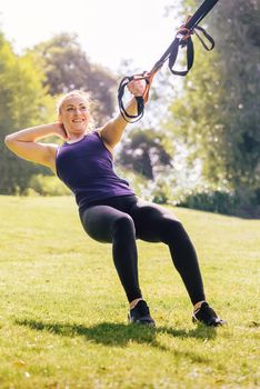 vertical photo of an athletic fitness woman that smiles training with suspension straps, during functional workout in a sunny outdoor park in the morning
