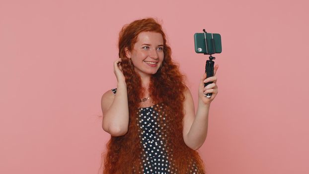 Redhead woman in dress traveler blogger taking selfie on mobile phone selfie stick, communicating video call online with subscribers. Young ginger girl isolated alone on pink studio background
