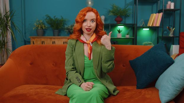 Great job. Redhead young woman in green dress showing thumbs up and nodding in approval, successful good work, satisfaction results at modern home apartment. Ginger girl in living room sitting on sofa