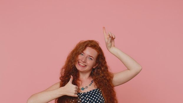 Lovely redhead woman showing thumbs up and pointing up empty place, advertising area for commercial text, copy-space for goods promotion. Young ginger girl. Indoors studio shot on pink wall background