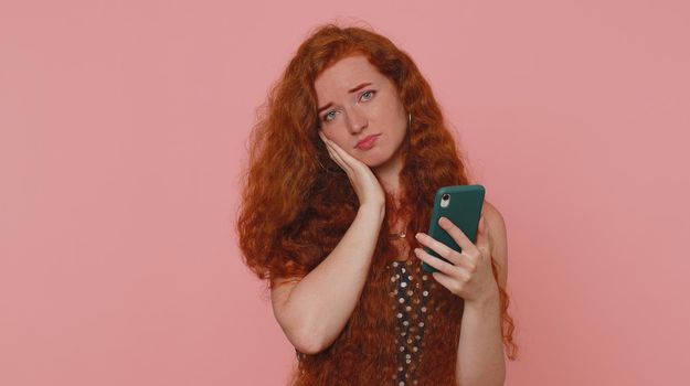 Unhappy sad young redhead woman use smartphone typing browsing, loses becoming surprised sudden lottery results, bad fortune, loss, unlucky news. Ginger girl with freckles on pink studio background