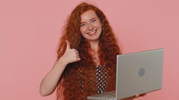 Redhead young woman working online on laptop finishing work, showing thumb up to camera, positive feedback, like, lottery win, approve good offer advertisement. Ginger girl on pink studio background