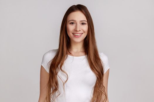 Portrait of beautiful woman with kind look and charming smile, standing and looking at camera, expressing positive emotions, wearing white T-shirt. Indoor studio shot isolated on gray background.