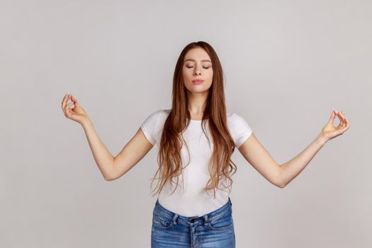 Portrait of calm beautiful woman holding hands up in mudra gesture, keeping eyes closed while meditating, yoga practice, wearing white T-shirt. Indoor studio shot isolated on gray background.