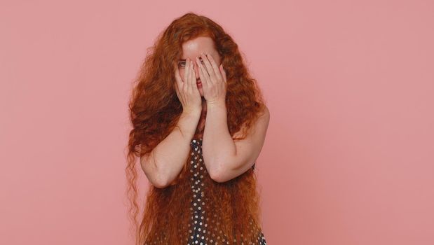 Nosy curious young redhead woman closing eyes with hand and spying through fingers, hiding and peeping, binocular gesture, exploring way, seeking something in distance. Ginger girl on pink background