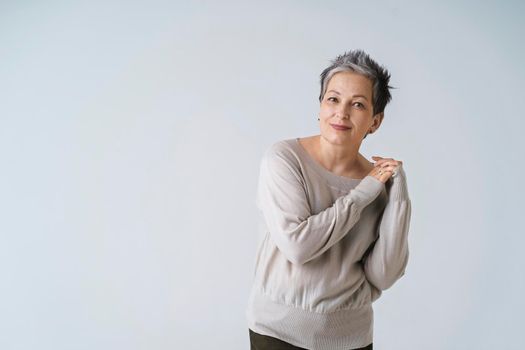 Smiling mature grey short hair woman posing with hands folded at her shoulder looking at camera wearing white blouse, copy space isolated on white background. Healthcare, aged beauty concept.