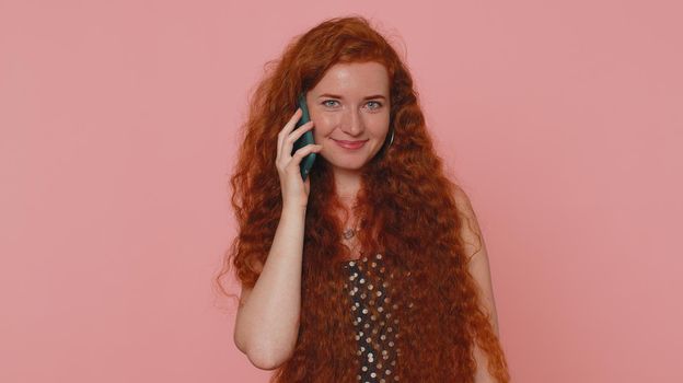Redhead young woman in polkadot dress having pleasant mobile conversation using smartphone with friends, telling good news. Adult ginger freckles face girl indoors isolated on pink studio background