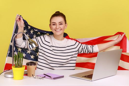 Happy woman raising American flag sitting at workplace and shouting for joy celebrating US Independence day 4th of july. Indoor studio studio shot isolated on yellow background.