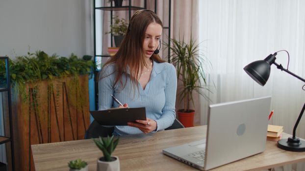Young business woman sitting at desk at home office wearing headset, freelance worker, call center, support service operator helpline, having talk with client or colleague at laptop computer notebook