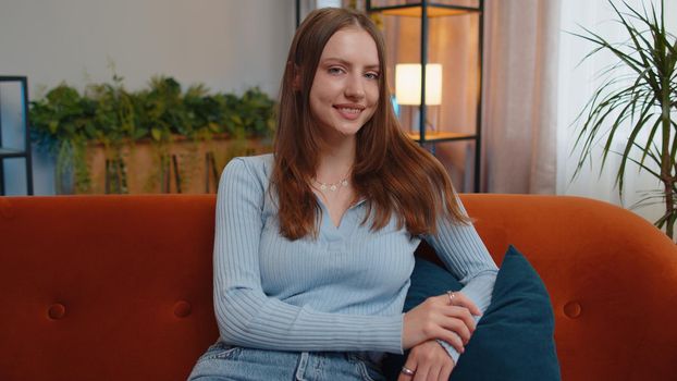 Close-up portrait of happy smiling young woman in blue blouse, looking at camera. Pretty beautiful brunette girl indoors isolated at home in living room sitting on couch alone. Female nature beauty