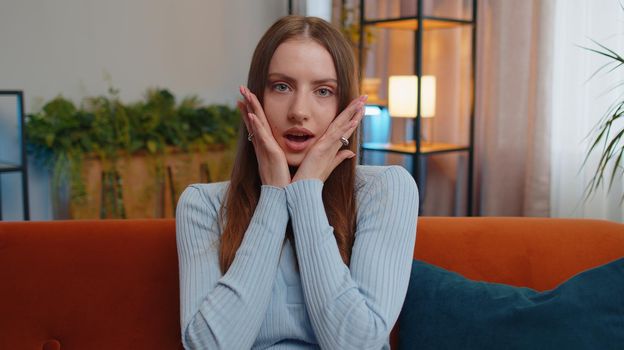 Oh my God, Wow. Young woman surprised looking at camera with big eyes, shocked by sudden victory, good win news, celebrating at home. Excited amazed pretty adult girl indoors in living room on couch