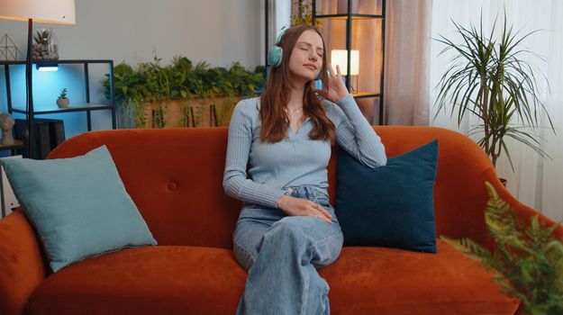 Happy relaxed overjoyed young woman in wireless headphones dancing on couch at home choosing listening favorite energetic disco rock n roll music in smartphone entertaining. People weekend activities