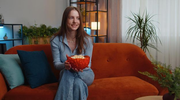 Excited young pretty woman sitting on sofa eating popcorn and watching interesting tv serial, sport game, film, online social media movie content online at home. Girl enjoying domestic entertainment