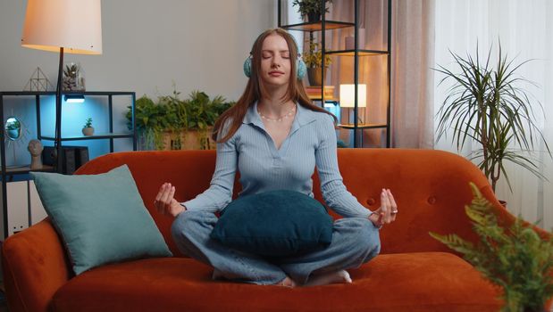 Keep calm down, relax, inner balance. Young woman breathes deeply with mudra gesture, eyes closed meditating with concentrated thoughts, peaceful mind. Tired girl sit at home in living room on couch