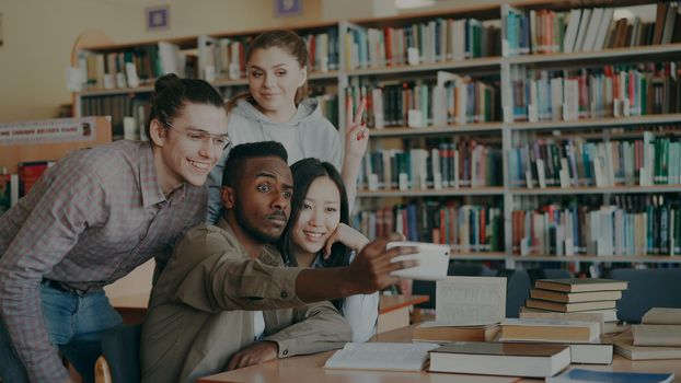 Group of international students have fun smiling and making selfie photos on smartphone camera at university library indoors. Cheerful friends have rest while prepare for examination