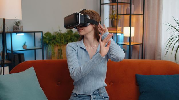 Young woman using virtual reality futuristic technology VR app headset helmet to play simulation 3D 360 video game, watching film movie at modern home apartment. Pretty girl in goggles sitting on sofa