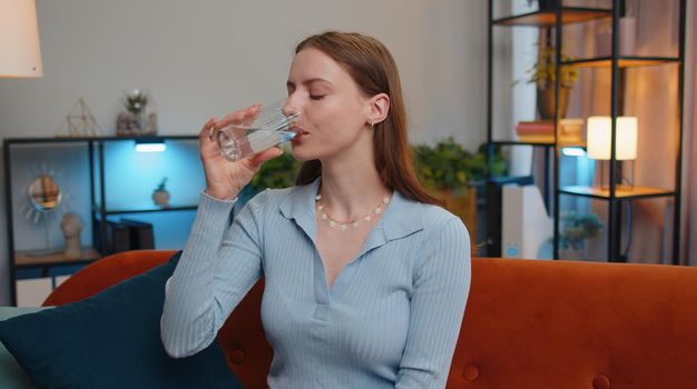 Portrait of thirsty young woman sitting indoors holding glass of natural aqua make sips drinking still water preventing dehydration. Girl with good life habits, healthy slimming, weight loss concept