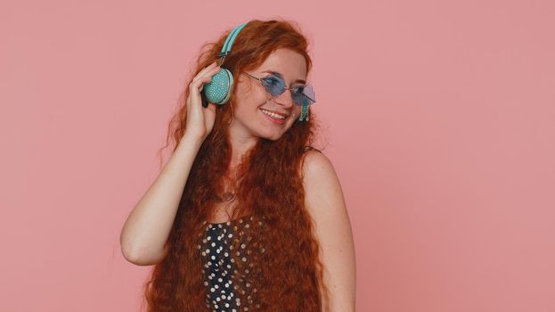 Young redhead woman in dress listening music on headphones, dancing disco fooling around having fun expressive gesticulating hands. Ginger girl with freckles isolated alone on pink studio background