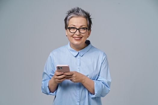 Texting or typing holding smartphone in hands mature grey haired woman working or shopping online, checking on social media. Pretty woman in blue blouse isolated on white background.