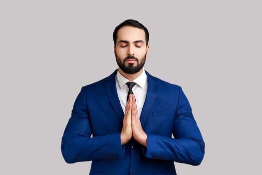 Portrait of calm handsome bearded businessman standing in yoga pose and try to relaxing, keeps palms together, wearing official style suit. Indoor studio shot isolated on gray background.