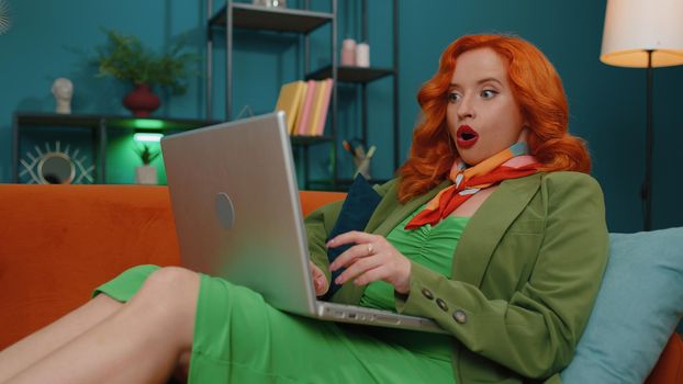 Oh my God Wow. Amazed redhead young woman use laptop computer, receive good news message, shocked by sudden victory, celebrate win, purchases online shopping at home. Ginger girl on living room couch