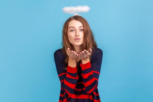 Kind woman wearing striped casual style sweater and nimb over head, sending air kiss over palms, expressing fondness, romantic feelings. Indoor studio shot isolated on blue background.