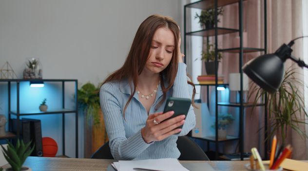 Young business woman use smartphone typing browsing, loses surprised by sudden lottery results, bad news, fortune, loss sitting at table working from home office. Girl freelancer remote job online