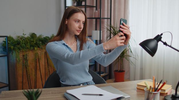 Young woman blogger taking selfie portraits on smartphone for social media vlog while sitting at table working from home office. Girl businesswoman freelancer study, e-learning, remote job online