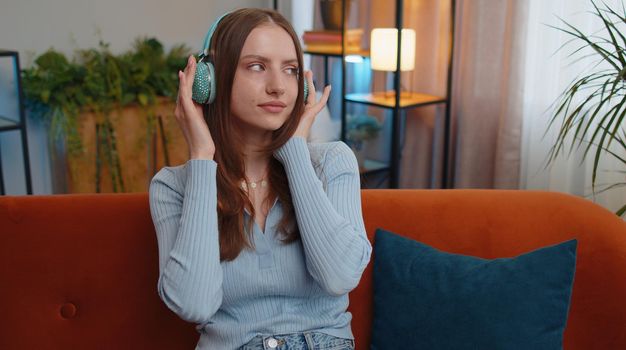 Portrait of happy relaxed overjoyed young woman in wireless headphones dancing on couch at home choosing listening favorite energetic disco music in smartphone entertaining. People weekend activities