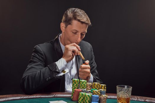 Arrogant male in a black slassic suit and white shirt is playing poker sitting at the table at casino in smoke, against a white spotlight. He rejoicing his victory smoking a cigar. Gambling addiction. Sincere emotions and entertainment concept.