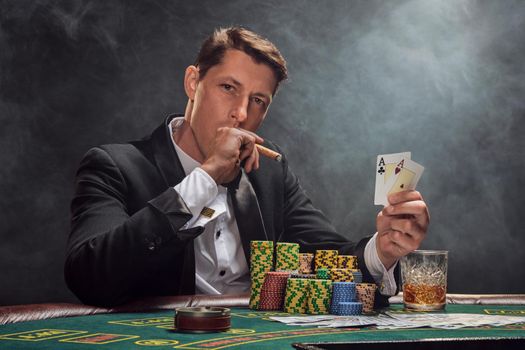 Smart man in a black slassic suit and white shirt is playing poker sitting at the table at casino in smoke, against a white spotlight. He rejoicing his victory showing two aces in his hand, smoking a cigar and looking at the camera. Gambling addiction. Sincere emotions and entertainment concept.