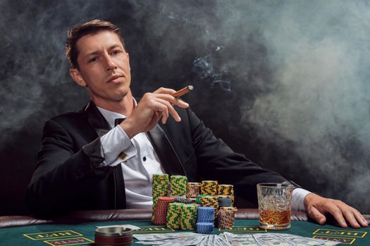 Smart person in a black slassic suit and white shirt is playing poker sitting at the table at casino in smoke, against a white spotlight. He rejoicing his victory smoking a cigar and looking at the camera. Gambling addiction. Sincere emotions and entertainment concept.