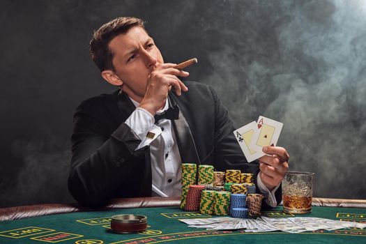 Elegant fellow in a black slassic suit and white shirt is playing poker sitting at the table at casino in smoke, against a white spotlight. He rejoicing his victory showing two aces in his hand and smoking a cigar. Gambling addiction. Sincere emotions and entertainment concept.