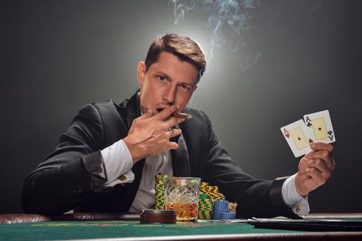 Good-looking person in a black slassic suit and white shirt is playing poker sitting at the table at casino in smoke, against a white spotlight. He rejoicing his win showing two aces in his hand, smoking a cigar and looking at the camera. Gambling addiction. Sincere emotions and entertainment concept.