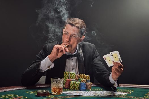 Good-looking fellow in a black slassic suit and white shirt is playing poker sitting at the table at casino in smoke, against a white spotlight. He rejoicing his victory showing two aces in his hand, smoking a cigar and looking away. Gambling addiction. Sincere emotions and entertainment concept.
