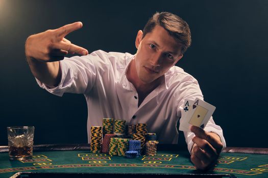 Handsome male in a white shirt is playing poker sitting at the table at casino in smoke, against a white spotlight. He rejoicing his victory holding two aces in his hand and showing a peace sign. Gambling addiction. Sincere emotions and entertainment concept.