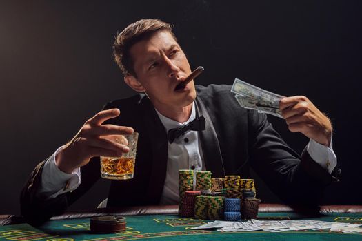 Handsome male in a black slassic suit and white shirt is playing poker sitting at the table at casino in smoke, against a white spotlight. He rejoicing his victory while smoking a cigar, drinking alcohol and holding some money in his hand. Gambling addiction. Sincere emotions and entertainment concept.