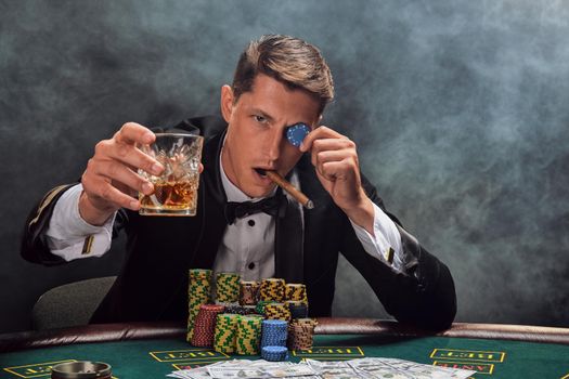 Stylish person in a black slassic suit and white shirt is playing poker sitting at the table at casino in smoke, against a white spotlight. He rejoicing his victory while drinking whiskey, smoking a cigar, fooling with a chip and looking at the camera. Gambling addiction. Sincere emotions and entertainment concept.