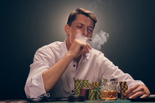 Elegant male in a white shirt is playing poker sitting at the table at casino in smoke, against a white spotlight. He rejoicing his victory and smoking a cigar. Gambling addiction. Sincere emotions and entertainment concept.