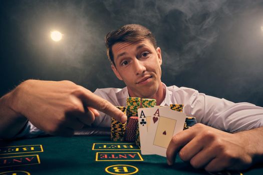 Close-up shot of a smart person in a white shirt is playing poker sitting at the table at casino in smoke, against a white spotlight. He rejoicing his victory showing two aces in his hand. Gambling addiction. Sincere emotions and entertainment concept.