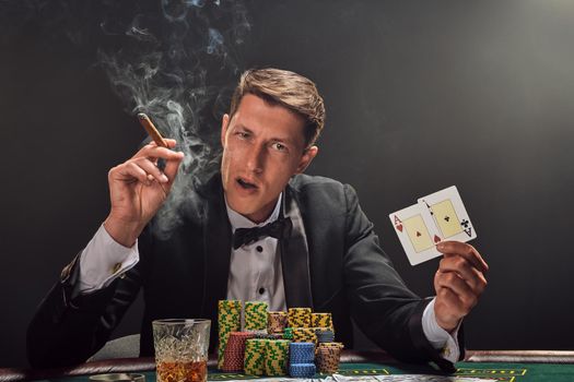 Good-looking guy in a black slassic suit and white shirt is playing poker sitting at the table at casino in smoke, against a white spotlight. He rejoicing his win showing two aces in his hand, smoking a cigar and looking at the camera. Gambling addiction. Sincere emotions and entertainment concept.