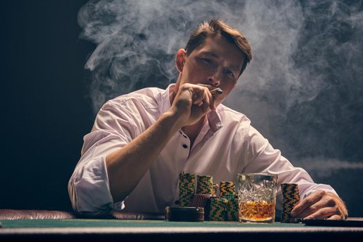 Handsome person in a white shirt is playing poker sitting at the table at casino in smoke, against a white spotlight. He rejoicing his victory and smoking a cigar. Gambling addiction. Sincere emotions and entertainment concept.