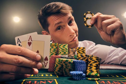 Close-up shot of a good-looking guy in a white shirt is playing poker sitting at the table at casino in smoke, against a white spotlight. He rejoicing his victory showing two aces and chips in his hands. Gambling addiction. Sincere emotions and entertainment concept.
