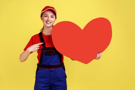 Smiling happy worker woman pointing at big red heart with finger, looking at camera, expressing positive emotions, wearing overalls and red cap. Indoor studio shot isolated on yellow background.