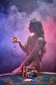 Portrait of a charming brunette girl with bright make-up, dressed in a golden glittered dress, posing sideways against a gambling table with a glass of champagne in her hand. Poker concept on a black smoke background with pink and blue backlights. Casino.