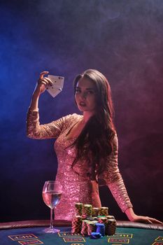 Portrait of an attractive brunette lady with bright make-up, dressed in a golden glittered dress, posing against a gambling table with two aces in her hands and looking at the camera. Poker concept on a black smoke background with pink and blue backlights. Casino.