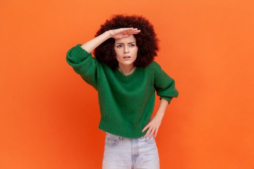 Attentive woman with Afro hairstyle wearing green casual sweater standing with hand near head, looking far away, trying to see something, bad vision.Indoor studio shot isolated on orange background.