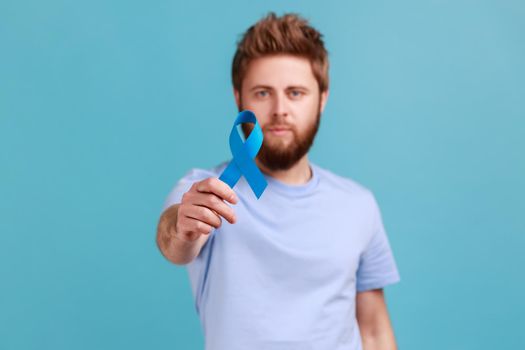 Portrait of serious young adult attractive bearded man holding out blue awareness, disease symbol, looking at camera, support and care. Indoor studio shot isolated on blue background.