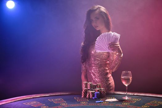 Portrait of a sexy brunette girl with bright make-up, dressed in a golden glittered dress, posing against a gambling table with a fan of hundred dollar bills in her hands and looking at the camera. Poker concept on a black smoke background with pink and blue backlights. Casino.