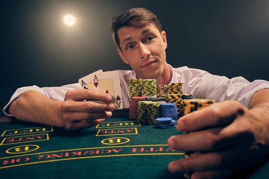 Close-up shot of a smart male in a white shirt is playing poker sitting at the table at casino in smoke, against a white spotlight. He rejoicing his victory showing two aces and some chips in his hands. Gambling addiction. Sincere emotions and entertainment concept.